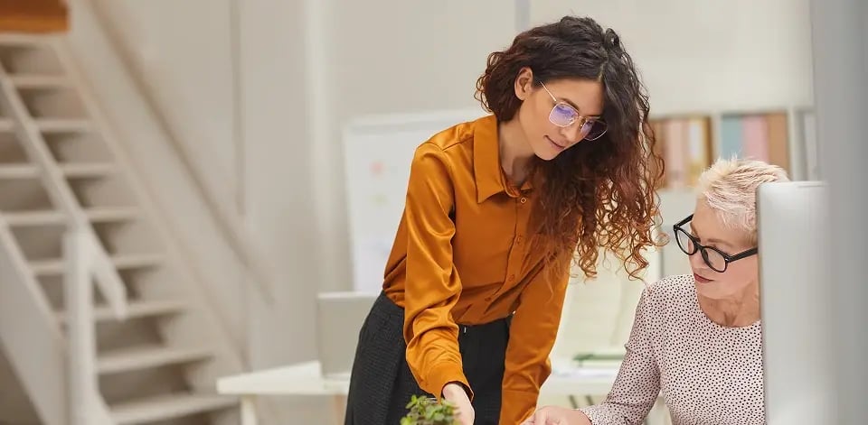 Two professional women collaborating at office desk reviewing documents in bright modern workspace