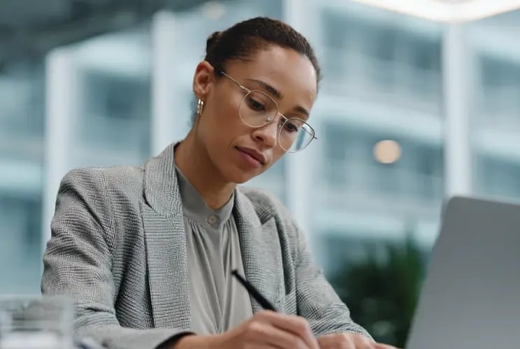 Professional woman in business attire writing notes while reviewing compliance documentation at office desk