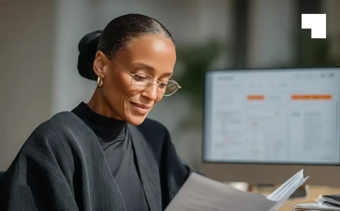 Professional woman in glasses reviewing documents at desk with computer monitor in background