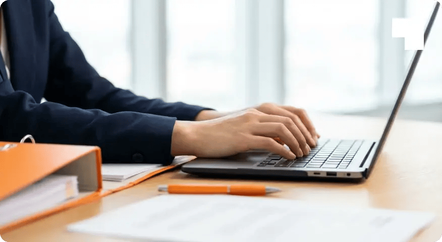 Close-up of hands mid-typing on a laptop keyboard at a bright modern office desk, orange binder and pen visible beside the laptop, clean bright white oak surface, no screen visible